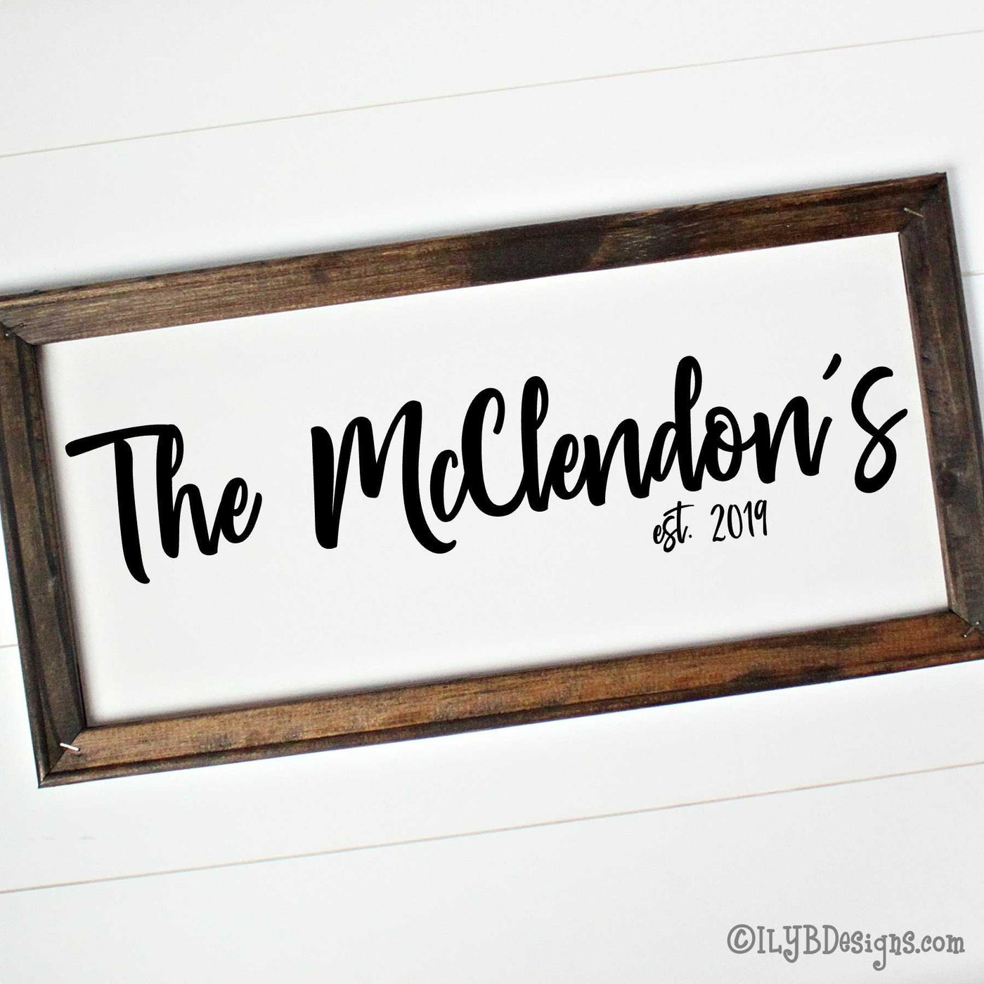 Dark walnut stained frame on a white canvas with a black design. Design is placed horizontally on the 20"x10" sign. Family name is in a bold script font with an established year underneath.