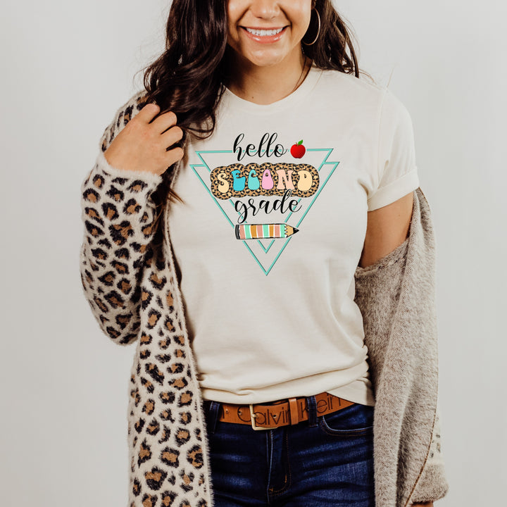 Woman wearing natural color grade level team teacher shirt featuring colorful retro letters with leopard print behind them and says hello second grade along with a pencil and apple.