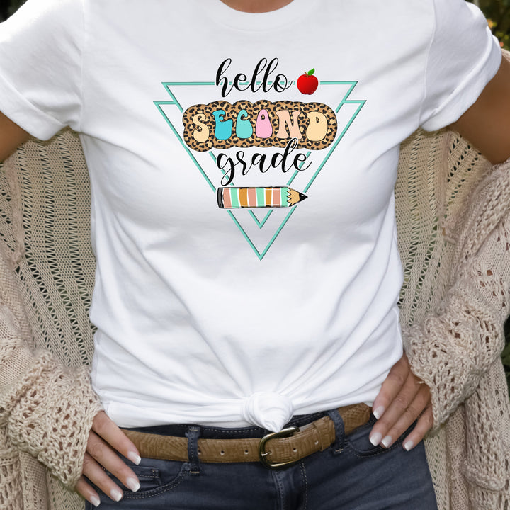 Woman wearing white grade level team teacher shirt featuring colorful retro letters with leopard print behind them and says hello second grade along with a pencil and apple.