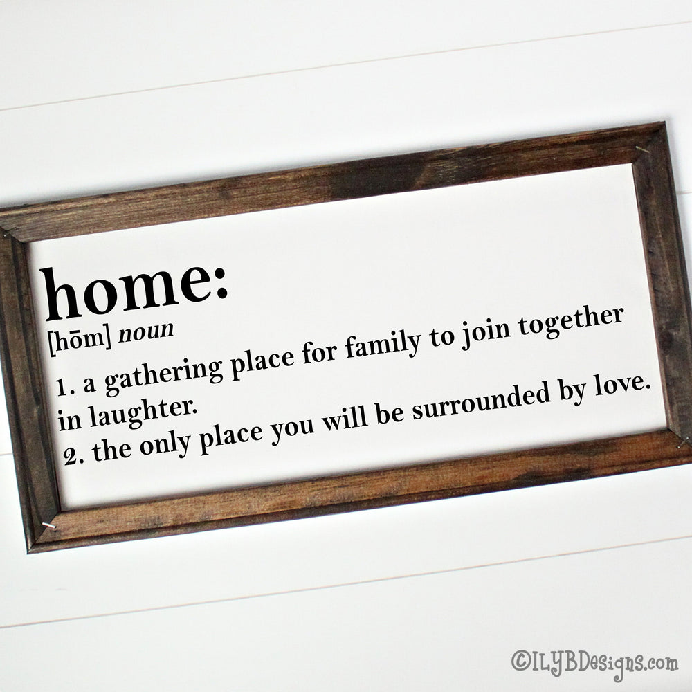 Dark walnut stained 20"x10" frame on a white canvas with black printed words that look like a dictionary definition.  It reads, "home: noun, 1. a gathering place for family to join together in laughter. 2. the only place you will be surrounded by love." Design is placed horizontally on the sign.