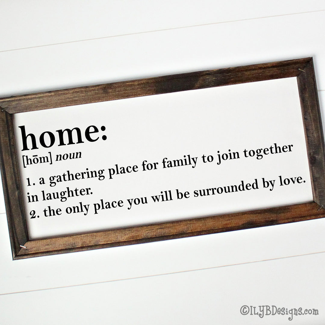 Dark walnut stained 20"x10" frame on a white canvas with black printed words that look like a dictionary definition.  It reads, "home: noun, 1. a gathering place for family to join together in laughter. 2. the only place you will be surrounded by love." Design is placed horizontally on the sign.