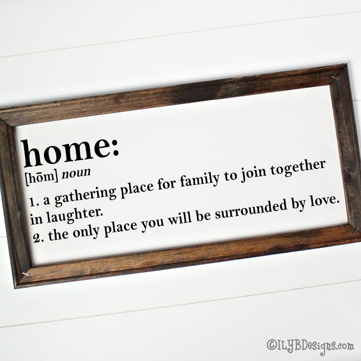 Dark walnut stained 20"x10" frame on a white canvas with black printed words that look like a dictionary definition.  It reads, "home: noun, 1. a gathering place for family to join together in laughter. 2. the only place you will be surrounded by love." Design is placed horizontally on the sign.