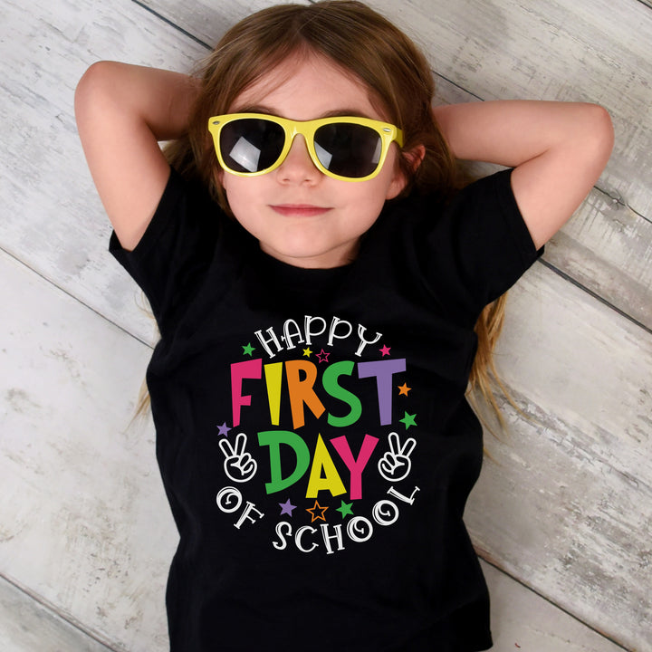 Young girl wearing a black shirt with a cheerful message in lively rainbow colored letters, spelling out Happy First Day of School in pink, yellow, orange, green, and purple letters.