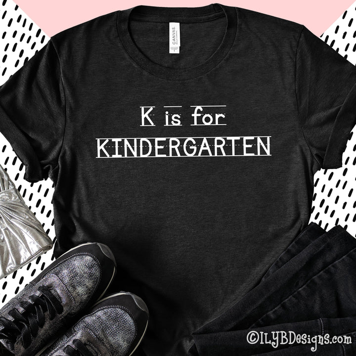 Heather black kindergarten teacher shirt that says K is for Kindergarten in a white font that looks like primary lined writing paper.