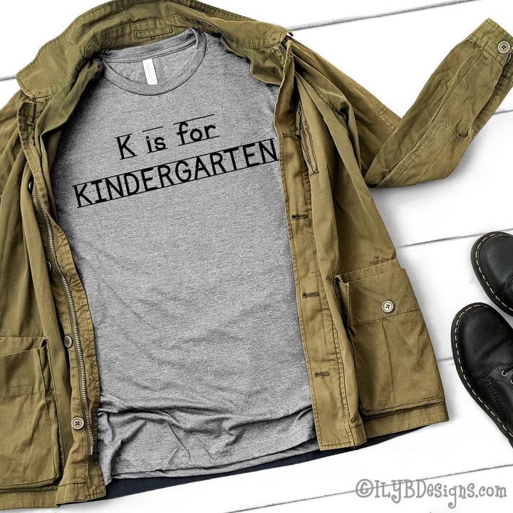 Athletic heather kindergarten teacher shirt that says K is for Kindergarten in a black font that looks like primary lined writing paper.