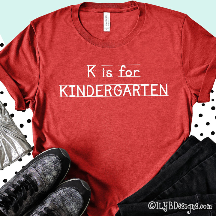 Heather red kindergarten teacher shirt that says K is for Kindergarten in a white font that looks like primary lined writing paper.