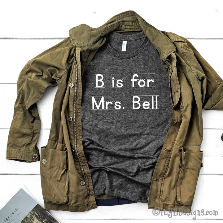 Dark heather gray personalized teacher shirt that says B is for Mrs. Bell in a white font that resembles primary lined writing paper.