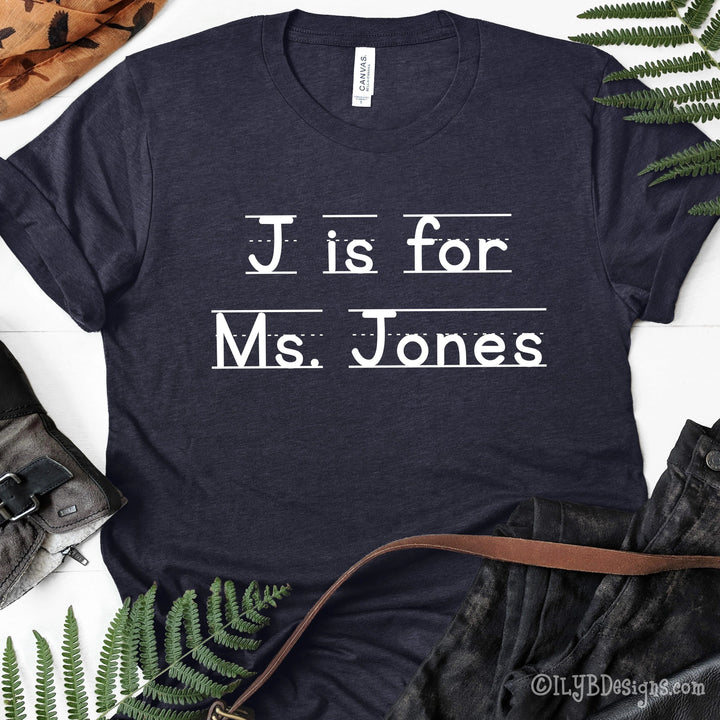 Heather navy personalized teacher shirt that says J is for Ms. Jones in a white font that resembles primary lined writing paper.