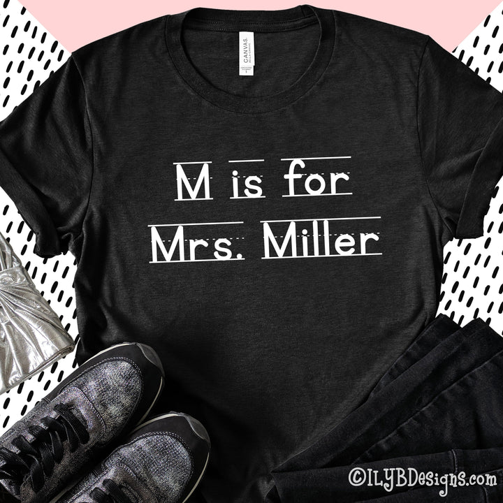 Heather black personalized teacher shirt that says M is for Mrs. Miller in a white font that resembles primary lined writing paper.