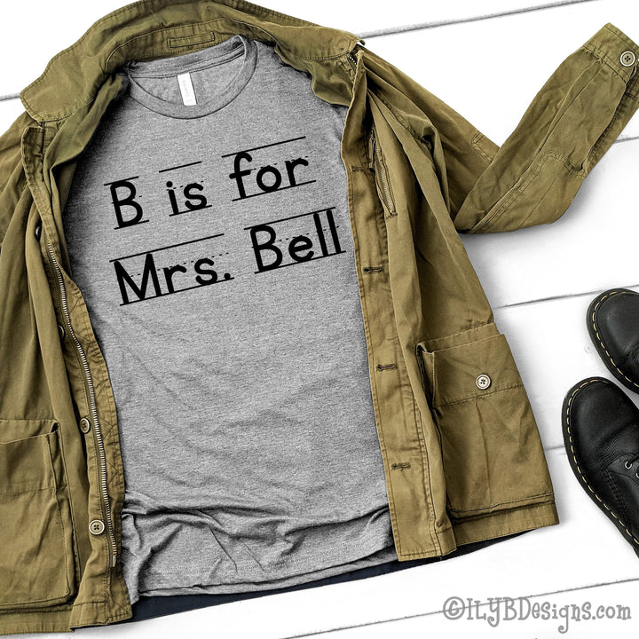 Athletic heather gray personalized teacher shirt that says B is for Mrs. Bell in a black font that resembles primary lined writing paper.