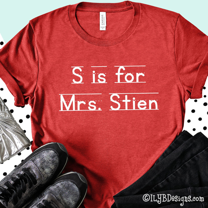 Heather red personalized teacher shirt that says S is for Mrs. Stien in a white font that resembles primary lined writing paper.
