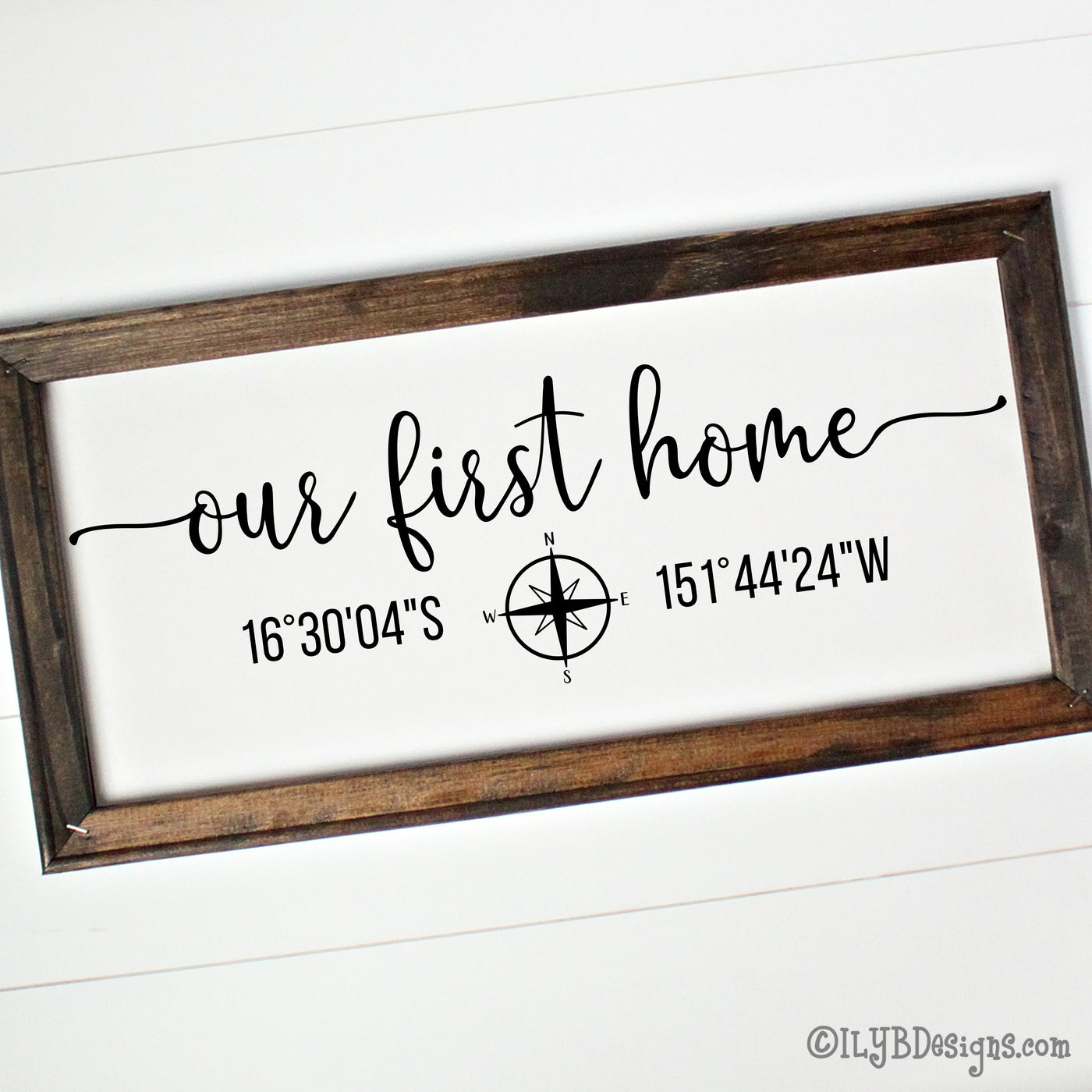 Our first home in a script font with a compass under the script and latitude and longitude coordinates on each side of the compass. Design is black on a white canvas and in a dark walnut stained wood frame.