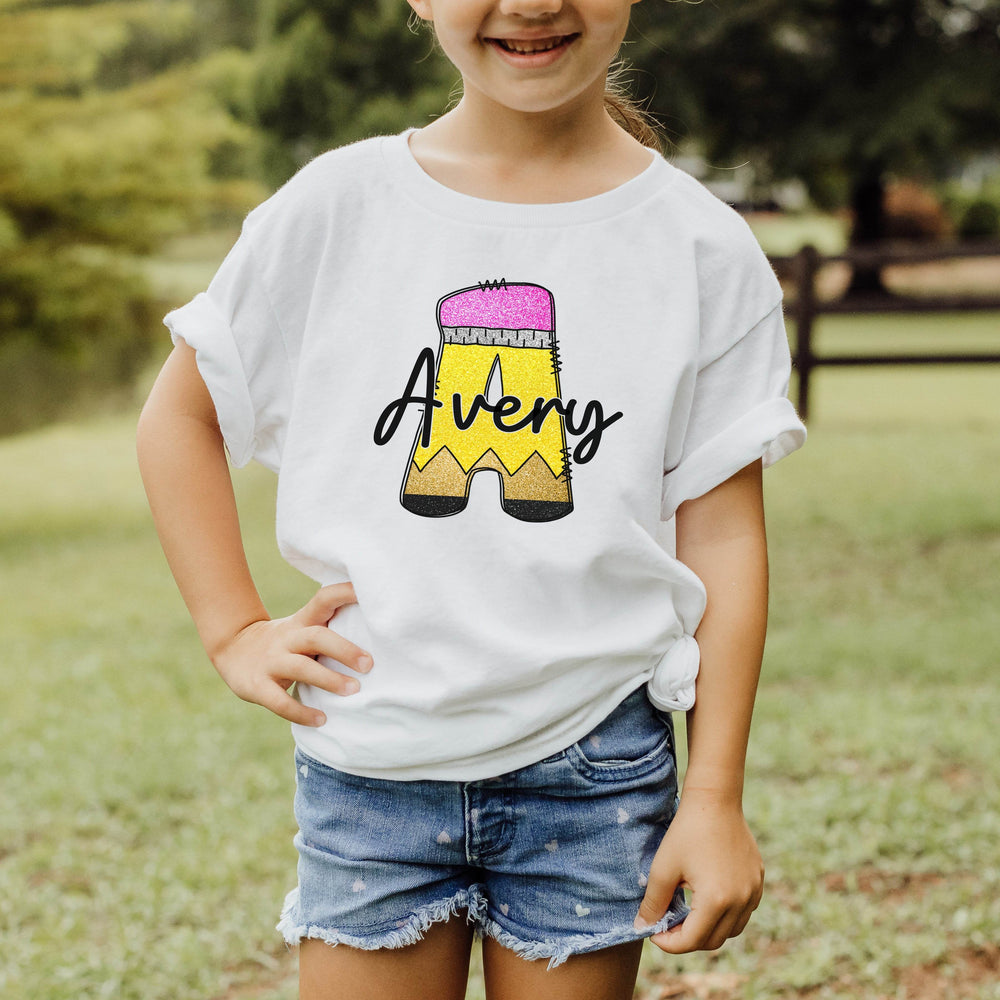 A young girl wearing a white t-shirt with a custom graphic of a yellow pencil and the name 'Avery' in black script, paired with denim shorts, standing outdoors in a grassy park.