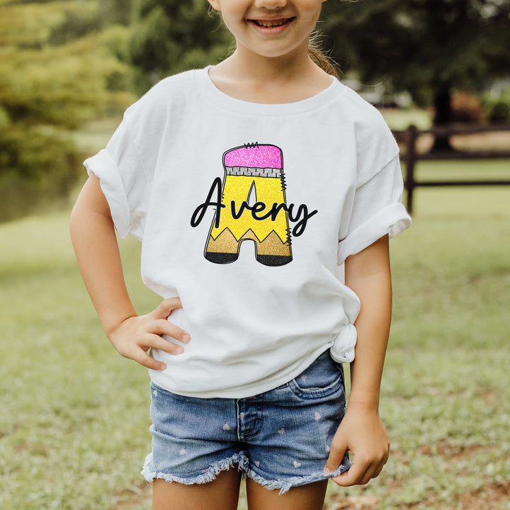 Young girl wearing a white personalized monogram school pencil back to school shirt with a large A that looks like a school pencil with a personalized name written in black script font across the middle of the pencil initial.