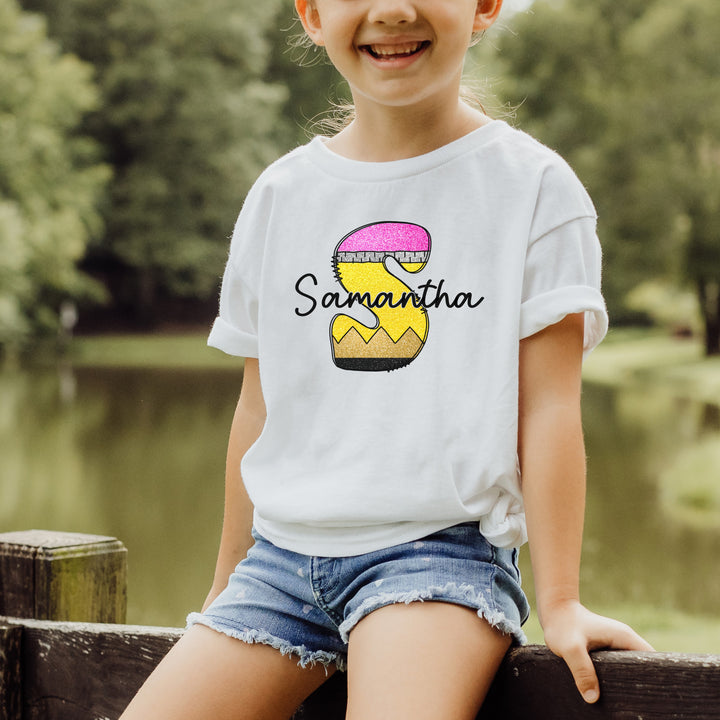 Young girl wearing a white personalized monogram school pencil back to school shirt with a large S that looks like a school pencil with a personalized name written in black script font across the middle of the pencil initial.
