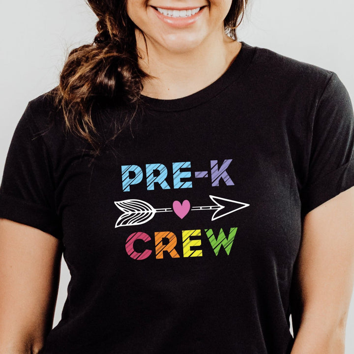Close up of woman wearing black personalized pre k crew shirt for preschool teachers featuring vibrant, rainbow colored letters spelling Pre-K Crew that pop against the dark fabric, creating a fun and playful look. An eye-catching arrow between the words adds an extra touch of whimsy, making this shirt a standout choice for any preschool teacher.  