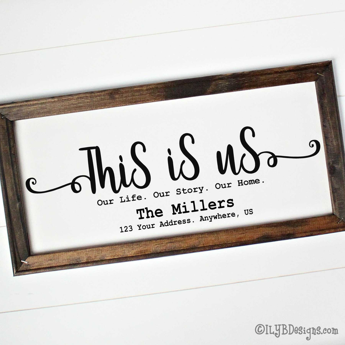 Dark walnut stained 20"x10" framed canvas with "This is us" in a black script font followed by a typewriter font that says, "Our Life. Our Story. Our Home." An optional family name is underneath those words. An optional address in under the family name.