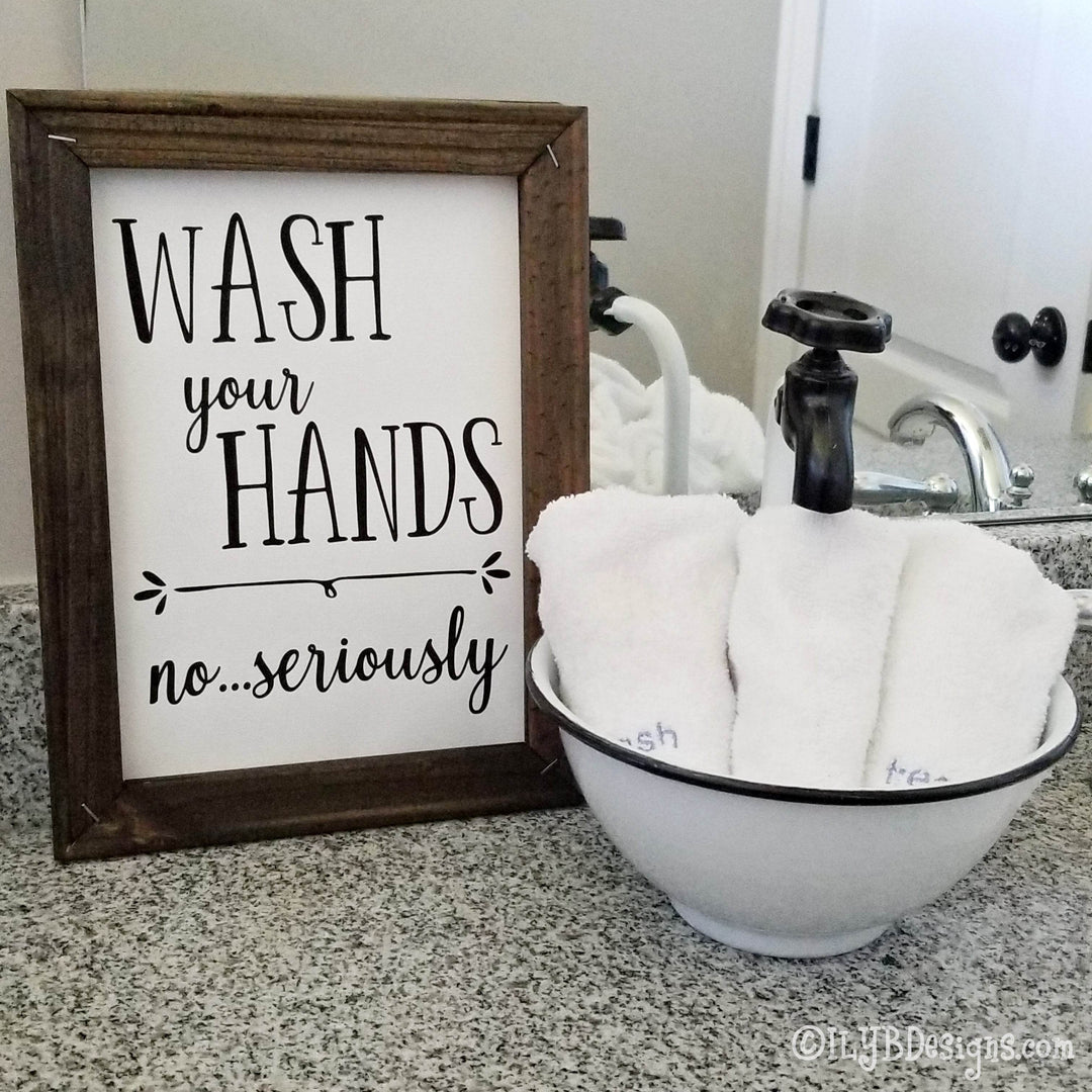 Framed canvas sign with 'Wash your hands no...seriously' black text on a white canvas background with a brown frame sitting on a bathroom counter.