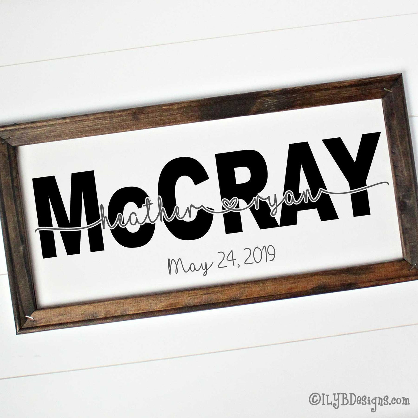 A dark walnut stained 20"x10" frame on a white canvas with a large, black last name in a bold print font. 2 first names in a silver gray script connected with an open heart are placed on the last name. An optional wedding date is below the name design in silver gray.